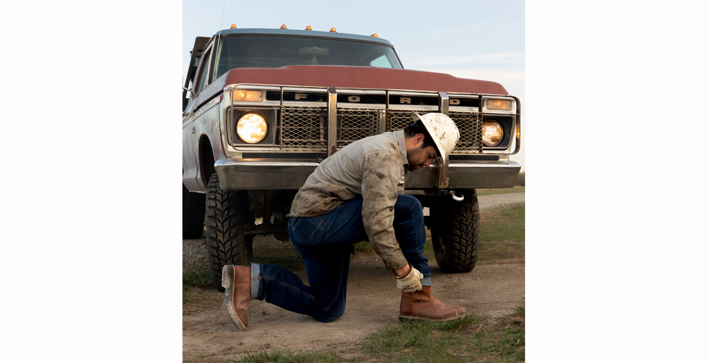 Un hombre posando delante de una vieja camioneta Ford y poniéndose las botas.